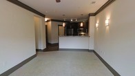 This single-floor space showcases a thoughtfully designed open-concept layout that seamlessly connects the living area to the kitchen. The kitchen is a standout feature, highlighted by dark espresso cabinetry, stainless steel appliances including a refrigerator and microwave, and a granite-topped peninsula that serves as a natural divider between the kitchen and living space. Warm red pendant lights hang above the peninsula, adding a pop of color and creating an inviting ambiance. A track lighting system further illuminates the kitchen workspace. The living area is finished with plush, light-toned carpeting that adds comfort underfoot, while dark baseboards and crown molding provide a sharp contrast against the neutral walls, lending a polished, cohesive aesthetic throughout the space. Wall sconces add additional warm lighting accents. A hallway visible from the main living area leads to additional rooms, suggesting a well-organized and functional floor plan. The overall design balances modern finishes with a welcoming atmosphere, making it a practical and visually appealing space.