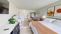 Located at 1057 Broward Rd in Jacksonville, Florida, this efficiently designed single-floor unit offers a well-utilized open layout that combines sleeping, lounging, and working areas into one cohesive space. The main room is generously proportioned, featuring light wood-style flooring, soft sage-green accent walls, and a textured ceiling that adds a classic touch. A large picture window floods the room with natural light and offers a pleasant view of the surrounding greenery, creating an airy and inviting atmosphere. The sleeping area is anchored by a king-sized bed and complemented by warm, earthy-toned wall art, adding personality and style to the space. A comfortable sofa, wooden coffee table, and an open metal shelving unit define the living area, while a dedicated workspace with a desk and wall-mounted TV provides functional versatility. A compact dining nook with a round white table completes the open floor plan. The bathroom features a full tub-shower combination wrapped in elegant marble-style tile with clean white fixtures and matte black accents, offering a crisp, modern aesthetic. This unit is thoughtfully arranged to maximize every square foot, making it a practical and stylish space in Jacksonville's 32218 area.