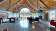 Located at 1941 Hidden Lake Drive in Stow, Ohio, this community lounge is a well-appointed gathering space designed to blend comfort, functionality, and modern aesthetics. The centerpiece of the main lounge area is a dramatic vaulted ceiling clad in warm natural wood planks, supported by white-painted structural beams that draw the eye upward. A floor-to-ceiling arched window wall floods the space with abundant natural light and frames sweeping views of the surrounding lush greenery and ornamental grasses. Polished concrete floors reflect the light throughout, enhancing the open, airy feel of the room.

A full-height stone fireplace anchors one wall, accompanied by a wall-mounted flat-screen TV and a seating arrangement of rich leather armchairs centered around glass coffee tables. A billiards table and shuffleboard offer additional recreational options. A dedicated business center features a vibrant, artistic mural backdrop, built-in workstations with computers, and casual seating with garden views.

The fully equipped kitchen showcases warm wood cabinetry, sleek grey countertops, stainless steel appliances, and an apron-front sink — a practical and attractive space for community gatherings. Cozy café-style nook seating with views of the landscaped grounds rounds out this inviting and versatile lounge.