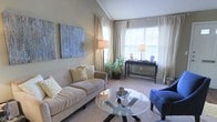 This single-story space centers around a thoughtfully designed living area that combines comfort with modern style. The room features vaulted ceilings that enhance the sense of openness, while large windows dressed in sheer, floor-length curtains allow abundant natural light to fill the space, creating a warm and airy atmosphere. The neutral color palette—comprising soft beiges and warm grays on the walls and carpet—provides a calm, cohesive backdrop throughout.

The living area is furnished with a plush, taupe sofa accented by yellow patterned throw pillows, complemented by a bold blue accent chair that adds a pop of color. A circular glass-top coffee table serves as a central focal point, contributing to the room's open feel. A tall floor lamp and a table lamp on a dark wood console table provide layered lighting options, while a large abstract canvas artwork on the wall adds a decorative statement. Lush indoor greenery near the window introduces a natural element, further enhancing the inviting ambiance of the space.