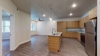 This single-floor unit offers a thoughtfully designed open-concept layout that seamlessly connects the kitchen and living area. The kitchen is a standout feature, showcasing flat-panel wood cabinetry in a warm natural tone, granite countertops, and a central island with an integrated sink and matching granite surface. Sleek pendant lights hang above the island, complementing the recessed lighting throughout the space. Stainless steel appliances, including a refrigerator, range, and microwave, complete the kitchen's modern aesthetic. The living area extends naturally from the kitchen, featuring durable wood-look plank flooring, neutral walls, and a ceiling fan that adds both function and comfort. Natural light filters in through windows, enhancing the open and airy feel of the space. The bedroom is carpeted for comfort, offering a quiet retreat with a window that brings in additional natural light and architectural ceiling detail that adds visual interest. The bathroom is clean and functional, featuring wood-look plank flooring, a combined tub and shower with a curved curtain rod, and a vanity with matching wood cabinetry. The overall design maintains a consistent modern aesthetic with warm wood tones, neutral finishes, and practical yet attractive fixtures throughout.