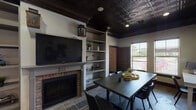 Located in Houston, TX (77040), this single-floor business center combines functionality with sophisticated design. The space is anchored by a large conference table surrounded by contemporary chairs, making it well-suited for meetings and collaborative work sessions. A dedicated computer workstation with a wall-mounted desk provides an additional productivity area along the perimeter.

A standout feature is the decorative brick fireplace, topped with a wall-mounted flat-screen TV, adding warmth and character to the room. Adjacent to the fireplace, built-in shelving displays curated décor, plants, and accessories. Comfortable leather lounge chairs and a round coffee table create an inviting seating nook ideal for informal conversations.

The dark, embossed tin ceiling with recessed lighting lends the space a distinctive, upscale aesthetic, complemented by rich dark wood trim and moldings throughout. Natural light filters in through multiple windows dressed with horizontal blinds, offering views of the surrounding exterior. The flooring transitions between wood-look planks and slate tile near the fireplace, adding visual texture. Abstract artwork on the walls and pops of yellow in the décor contribute a modern, energetic touch to the overall ambiance. A French-door entry connects seamlessly to adjacent rooms.