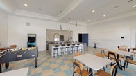 This single-floor community gathering space blends playful design with functional amenities, creating an inviting environment for socializing and recreation. The signature blue, cream, and yellow checkered tile flooring ties the entire space together with a retro-inspired aesthetic, while bright white walls and tray ceilings with recessed lighting give the room an airy, open feel.

At the heart of the space is a central island bar with sleek countertops and black bar stools, ideal for casual dining or socializing. The adjacent kitchenette alcove is finished with glossy white subway tile, shiplap walls, white cabinetry, stainless steel appliances including a beverage cooler, and a sink — perfectly equipped for hosting events.

The dining area features multiple tables with bentwood chairs arranged to accommodate both small and large groups, alongside wall-mounted TVs for entertainment. A large corkboard display wall and a foosball table anchor the game zone, which also includes a classic arcade cabinet, adding a fun, recreational atmosphere.

Expansive glass sliding doors and large windows flood the space with natural light and offer direct views of an outdoor patio area featuring palm trees and shaded seating with yellow umbrellas, seamlessly connecting the interior to the exterior. Wainscoting details further elevate the space's polished, community-focused character.