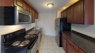 This single-floor residence offers a thoughtfully arranged layout across several distinct living areas. The spacious main living room features warm hardwood-style flooring, crown molding, and a charming built-in wood shelving unit set into the wall, adding a classic architectural touch. Large double-hung windows flood the room with natural light and provide views of the surrounding urban streetscape, while baseboard heating units ensure year-round comfort. An open entryway connects the living room to the rest of the unit, creating a seamless flow throughout the space. The kitchen is well-appointed with rich dark wood cabinetry, dark granite-style countertops, and a full suite of stainless steel appliances including a refrigerator, electric range, and over-the-range microwave. Light ceramic tile flooring completes the kitchen's practical design. The bedrooms continue the hardwood-style flooring and feature drop tile ceilings, neutral gray and white walls, and ample natural light from large windows. Closet access is present in at least one bedroom. The bathroom retains a vintage character with warm terracotta-toned wall tiles, a full soaking tub with shower, mosaic tile flooring, and a window for natural ventilation. Throughout the space, clean white trim and neutral tones create a cohesive and inviting atmosphere.