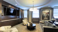 This refined single-floor space exudes understated elegance through a thoughtful combination of architectural details and upscale finishes. The centerpiece of the main lounge area is a striking circular tray ceiling, which frames a large drum pendant light suspended from a polished chrome fixture, casting a warm and inviting glow throughout the room. Rich, dark wood wall paneling lines the walls, lending depth and a sense of luxury to the interior, while a wall-mounted flat-screen television is seamlessly integrated into the built-in cabinetry for a clean, streamlined look. Natural light filters generously into the space through multiple windows dressed with soft, layered drapery in neutral tones, creating a bright yet comfortable atmosphere. The seating arrangement includes a plush sofa, a classic floral accent chair, and an upholstered ottoman, all centered around a sleek, dark round coffee table — a layout that encourages relaxed conversation. Decorative artwork adds a finishing touch to the walls, complementing the space's warm color palette of taupe, cream, and espresso tones. The flooring features a light tile or stone pattern that contrasts elegantly with the darker furnishings. Overall, the space presents a polished, welcoming ambiance well-suited for both leisure and formal gatherings.