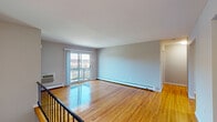 This single-floor residence presents a thoughtfully laid-out living space defined by its warm, polished hardwood floors that run throughout. The open living area is a standout feature, bathed in natural light streaming through large sliding glass doors that lead directly to a private balcony with outdoor views. A wall-unit air conditioner and baseboard heating ensure year-round comfort. The neutral gray wall tones throughout the home create a calm, cohesive aesthetic that complements the richness of the hardwood flooring.

The bedrooms are generously proportioned, each featuring the same gleaming hardwood floors and baseboard heating. One bedroom includes a dedicated walk-in closet with built-in shelving, offering practical storage solutions. Windows with blinds provide both natural light and privacy in the sleeping areas.

The bathroom is a refined space, featuring a full soaking tub surrounded by clean white subway tiles accented with elegant tan and dark gray decorative border strips. A frosted window allows natural light while maintaining privacy, and dark floor tiles complete the polished look.

A staircase with a classic black metal railing connects this floor to the entry level, adding a structural design element to the overall layout. The space is clean, move-in ready, and offers a comfortable, functional living environment.