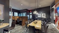 This single-floor co-working space is designed with a contemporary, urban aesthetic, combining dark charcoal walls, warm wood accents, and patterned carpeting throughout. The main conference area serves as the centerpiece, featuring a large natural wood communal table surrounded by tufted leather chairs, overhead industrial-style pendant lighting, a wall-mounted television, and a vibrant, Texas-themed graphic mural that energizes the space with bold typography and illustrative artwork.

Adjacent to the conference area, a lounge zone offers high-top bistro tables with bar stools, framed abstract artwork on dark walls, and access to a dedicated coffee bar — its entrance marked by a playful typographic wall installation. The coffee bar area includes dark cabinetry and counter space stocked for daily use.

A compact kitchenette is tucked nearby, finished with white subway brick tile, dark lower cabinets, a stainless steel under-counter refrigerator, a single basin sink, and a coffee maker — providing essential conveniences in a well-organized layout.

Private studio offices line one section of the space, enclosed with glass-paneled folding doors and labeled individually. Open workstations with rich wood panel dividers and ergonomic task chairs offer semi-private work areas with access to natural light through clerestory-style windows along the perimeter.