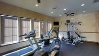 This dedicated fitness center offers a comprehensive workout environment designed to support a variety of exercise routines. The space is outfitted with multiple cardio machines, including treadmills, an elliptical trainer, and a recumbent stationary bike, all positioned along a row of windows fitted with warm wood-slat blinds that allow natural light to filter in while maintaining privacy. A multi-station cable weight machine anchors the strength training area, providing options for a full-body resistance workout. A wall-mounted flat-screen television is conveniently positioned for viewing during exercise sessions. The ceiling features recessed lighting alongside two ceiling fans, ensuring the space remains well-lit and comfortably ventilated during use. The walls are finished in a warm neutral tone with a textured appearance, contributing to a clean and inviting atmosphere. Dark flooring runs throughout, providing a practical and durable surface suited for fitness activities. A decorative potted plant adds a subtle natural element to the room. Overall, the fitness center is thoughtfully arranged to maximize usable floor space while keeping equipment accessible and the environment comfortable for daily workouts.