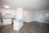 This single-floor residence offers a clean, contemporary layout designed for comfortable everyday living. The open-concept living and dining area is anchored by durable wood-look plank flooring in a warm gray tone, neutral walls, and a vertical blind-covered sliding door that allows natural light to fill the space. The kitchen is positioned adjacent to the living area and features white shaker-style cabinetry, granite-look countertops, stainless steel appliances including a dishwasher and range, and a stainless undermount sink — all set atop matching wood-look flooring for a cohesive aesthetic. A peninsula counter provides a clear visual separation between the kitchen and living space. The bedrooms are carpeted in a neutral plush carpet, offering a comfortable retreat, with one bedroom featuring a window that brings in natural light and outdoor views. The bathroom is outfitted with a full-size tub and shower combination, a white vanity with granite-look countertop, brushed nickel hardware, and wood-look flooring that ties back to the main living areas. Throughout the residence, a consistent neutral color palette and modern finishes create a cohesive and move-in ready environment.
