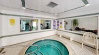 This single-level spa facility is designed with a clean, functional aesthetic centered around a large circular hot tub as its focal point. The interior is finished with white ceramic tile walls accented by horizontal green tile borders, creating a bright and well-maintained environment. The flooring is covered in small square tiles, practical and slip-resistant, extending throughout the space.

Large windows line one wall, offering views into the adjacent fitness center, which is visible equipped with cardio machines and weight equipment. This layout creates a connected wellness environment while keeping each area distinct. A glass-paneled door provides direct access to an outdoor area, allowing natural light to filter into the room and offering easy egress.

Seating is available along the perimeter with several plastic chairs and a small table, providing a comfortable resting area for guests. The ceiling features a paneled design with integrated ventilation and recessed lighting, keeping the atmosphere well-lit and properly ventilated. Pool rules and safety signage are posted prominently on the walls. Overall, the spa area offers a straightforward, well-equipped relaxation space suited for recreational use.