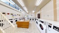 This shared laundry facility offers a practical and well-organized space across a single floor. The interior is finished with white square wall tiles accented by warm beige vertical strips, contributing to a clean and functional aesthetic throughout. Durable terrazzo flooring covers the entire area, providing a low-maintenance and visually cohesive surface underfoot.

The main laundry area is lined with multiple commercial-grade front-loading washing machines and Speed Queen commercial dryers arranged along the perimeter walls, offering ample capacity for high-volume use. A central wooden folding island with a built-in sink serves as a practical workstation for sorting and preparing laundry. Additional utility sinks are also integrated into the island's surface.

Bright fluorescent strip lighting runs across the ceiling, ensuring even illumination throughout the space. High clerestory windows along one wall allow natural light to filter in, enhancing the open and airy feel of the room. A staircase with a white metal railing is present in one section of the facility, indicating access to adjacent areas. Notice boards mounted on the tiled walls provide instructional and informational postings for users.

Decorative potted plants and hanging greenery are displayed on shelving above the machines, adding a welcoming touch to the otherwise utilitarian environment. The overall layout is spacious and thoughtfully arranged for efficient, comfortable use.