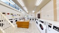 This shared laundry facility offers a practical and well-organized space across a single floor. The interior is finished with white square wall tiles accented by warm beige vertical strips, contributing to a clean and functional aesthetic throughout. Durable terrazzo flooring covers the entire area, providing a low-maintenance and visually cohesive surface underfoot.

The main laundry area is lined with multiple commercial-grade front-loading washing machines and Speed Queen commercial dryers arranged along the perimeter walls, offering ample capacity for high-volume use. A central wooden folding island with a built-in sink serves as a practical workstation for sorting and preparing laundry. Additional utility sinks are also integrated into the island's surface.

Bright fluorescent strip lighting runs across the ceiling, ensuring even illumination throughout the space. High clerestory windows along one wall allow natural light to filter in, enhancing the open and airy feel of the room. A staircase with a white metal railing is present in one section of the facility, indicating access to adjacent areas. Notice boards mounted on the tiled walls provide instructional and informational postings for users.

Decorative potted plants and hanging greenery are displayed on shelving above the machines, adding a welcoming touch to the otherwise utilitarian environment. The overall layout is spacious and thoughtfully arranged for efficient, comfortable use.