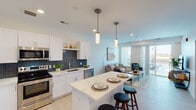 This thoughtfully designed single-floor residence offers a bright, open-concept layout that seamlessly connects the kitchen, dining, and living areas. The kitchen is a standout feature, outfitted with crisp white cabinetry, quartz countertops, a striking navy blue mosaic tile backsplash, and a full suite of stainless steel appliances including a refrigerator, range, microwave, and dishwasher. A large center island with seating provides both additional prep space and a casual dining option, while pendant lighting adds a polished finishing touch overhead.

The living area flows naturally from the kitchen and is bathed in natural light from large windows and a sliding glass door that opens to a private balcony, offering outdoor views of the surrounding neighborhood. Light hardwood-style flooring runs throughout the main living spaces, enhancing the airy, contemporary feel of the home.

The bedroom is a calm and comfortable retreat, featuring plush carpeting, a generously sized window with views to the outside, and clean neutral walls that create a serene atmosphere. The bathroom continues the modern aesthetic with large-format white subway tiles, a glass-enclosed walk-in shower, and a decorative mosaic tile accent strip that adds visual interest. Overall, the space presents a clean, cohesive design with quality finishes throughout.