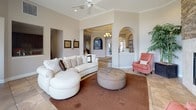 This refined single-floor community space offers a welcoming and sophisticated atmosphere from the moment you enter. The grand entry foyer greets visitors with a chandelier overhead, large-format tile flooring, decorative arched alcoves, and a neatly appointed console table — setting an elegant tone throughout. A central hallway connects the various areas seamlessly, anchored by a built-in media niche featuring a television and ornamental cabinetry that serves as an attractive focal point.

The spacious lounge area is a standout, featuring a dramatic floor-to-ceiling stacked stone fireplace framed by cream tile, complemented by plush seating including a curved white leather sofa, a tufted ottoman, and coral accent chairs. Tall windows dressed in rich chocolate drapery flood the space with natural light while offering views of lush landscaping outside.

A separate seating nook provides a more intimate setting with a glass-topped table and tufted chairs, ideal for private meetings or consultations. The kitchenette is outfitted with rich dark wood cabinetry, granite countertops, and a compact refrigerator — perfect for hosting refreshments. Throughout, crown molding, warm neutral walls, decorative artwork, and carefully chosen furnishings contribute to a polished, cohesive ambiance.