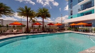 Located at 3300 Boutwell Rd in Lake Worth Beach, Florida, this outdoor pool area offers a vibrant, resort-style experience designed for relaxation and leisure. The centerpiece is a generously sized, crystal-clear swimming pool with a gradual depth, bordered by a tastefully paved deck finished in neutral stone tiles that complement the surrounding architecture.

Tall, swaying palm trees are positioned throughout the pool deck, providing natural shade and reinforcing the tropical Florida ambiance. Vivid orange market umbrellas are scattered across the space, sheltering a variety of seating arrangements — from lounge chairs ideal for sunbathing to bistro-style tables and chairs suited for casual outdoor dining or socializing.

The pool area is framed on multiple sides by a modern multi-story building, whose exterior is finished in a striking combination of sky blue, teal, and white — lending the space a fresh, contemporary coastal aesthetic. Ground-level sliding glass doors and upper-floor balconies connect the interior to this outdoor retreat, enhancing the open, airy feel.

Lush tropical plantings, including large potted palms and flowering plants, are thoughtfully integrated into the space, adding greenery and life to the hardscaped surroundings. The pool deck also includes practical amenities such as rinse stations and waste receptacles. Bathed in abundant South Florida sunshine, this outdoor area exudes a welcoming, resort-like atmosphere year-round.