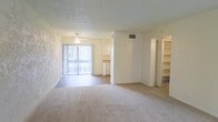 This single-floor residence offers a clean, open layout designed for comfortable everyday living. The main living area is carpeted and bathed in natural light, courtesy of a large sliding glass door fitted with vertical blinds that leads to an outdoor balcony or patio space. A ceiling fan is centrally positioned overhead, providing both lighting and air circulation, while a textured popcorn ceiling adds a classic architectural touch.

The open floor plan allows for a seamless transition between the living area and the adjoining spaces. To one side, a built-in shelving unit offers generous storage and display space, ideal for books, décor, or organizational needs. The adjacent area provides a glimpse into what appears to be a kitchen or utility nook, featuring light-colored cabinetry and countertop surfaces.

The walls carry a soft, neutral tone, complemented by light-colored carpeting that adds warmth underfoot. HVAC vents are integrated into the ceiling, ensuring climate control throughout. The overall ambiance is bright, airy, and move-in ready, with a straightforward layout that makes the space feel open and versatile. The design emphasizes practicality while maintaining a welcoming, light-filled atmosphere throughout.