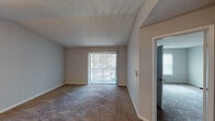 This single-story unit is thoughtfully laid out across several rooms, each finished with consistent, neutral-toned details. The main living area is generously sized, featuring wall-to-wall carpet in a warm beige tone, a textured ceiling, and a full-width sliding glass door fitted with vertical blinds that allow natural light to filter in while providing outdoor views. A smart thermostat is mounted on the wall, and the room connects seamlessly to adjoining spaces.

The bedrooms are carpeted and well-proportioned, each receiving natural light through windows dressed with horizontal blinds. At least one bedroom includes a built-in closet with a standard rod for hanging storage, while another features vaulted ceiling angles that add visual interest. The kitchen is equipped with white shaker-style cabinetry, light speckled countertops, a black electric range with a range hood above, and ample upper cabinet storage. The bathroom includes a white vanity with a single basin, light speckled countertop, a large mirror, vanity lighting, and dark hardwood-style flooring—offering a clean, functional finish throughout.