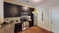 This single-floor residence at 1127 S Mint St in Charlotte's vibrant South End neighborhood offers a well-designed layout with contemporary finishes throughout. The open-concept living area is anchored by plush carpeting, a bold accent wall in deep charcoal gray, and a sliding glass door that leads to a private balcony overlooking the surrounding community. Black-and-white chevron drapery adds a graphic design element that carries cohesively through the space.

The kitchen is outfitted with dark espresso cabinetry extending to the ceiling for ample storage, stone-patterned countertops, and a full suite of stainless steel appliances including a range, microwave, and refrigerator. Hardwood-style flooring grounds the kitchen and flows through the adjacent areas, providing a warm contrast to the cabinetry.

The bedroom is generously proportioned, featuring soft carpeting, yellow-toned walls, and a bay of windows that flood the room with natural light and offer views of the surrounding greenery. A sitting area near the windows adds functional versatility to the space.

The bathroom is finished with tile flooring, a tiled tub-shower combination, a dark vanity with a white countertop, and bold drapery that echoes the home's consistent color palette of black, white, and yellow.