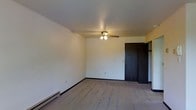 This single-floor space presents a straightforward, functional layout centered around a generously sized room visible in the image. The room features neutral, light-colored walls that contribute to a clean and understated aesthetic. Underfoot, wall-to-wall carpeting in a muted beige tone adds warmth and comfort to the space.

A ceiling fan with an integrated light fixture is centrally mounted, providing both air circulation and ambient lighting. Along the base of one wall, a baseboard heater offers a dedicated heating solution, a practical feature suited for colder climates.

A dark-framed door is positioned along one wall, providing access to an adjoining area that appears to include additional storage or closet space, partially visible through the open door. The dark trim of the door and baseboard creates a subtle contrast against the light walls, adding a modest degree of visual definition to the room.

The overall ambiance is simple and neutral, offering a blank canvas well-suited for a variety of uses. The space is move-in ready and designed with practical, everyday functionality in mind.