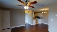 This single-story space offers an open-concept layout that seamlessly connects the living area to the kitchen, creating a cohesive and functional flow throughout. The living area features rich, dark hardwood flooring that extends throughout the space, complemented by neutral taupe-toned walls and clean white baseboards that add a crisp contrast. A five-blade ceiling fan with an integrated light fixture serves as a practical centerpiece overhead, providing both air circulation and ambient lighting.

The kitchen is efficiently designed with white cabinetry offering ample storage, dark countertops, and a range of standard appliances including a stove and dishwasher. A partial dividing wall separates the kitchen from the living area while maintaining the open feel of the space. An elegant chandelier above the dining area adds a refined touch and defines the transition between spaces.

A doorway visible from the living area leads to additional rooms, suggesting a well-organized floor plan. Ceiling vents throughout ensure consistent climate control. The combination of warm wood flooring, neutral walls, and functional fixtures gives the space a balanced, move-in-ready ambiance.
