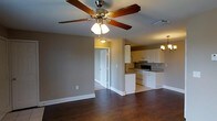 This single-story space offers an open-concept layout that seamlessly connects the living area to the kitchen, creating a cohesive and functional flow throughout. The living area features rich, dark hardwood flooring that extends throughout the space, complemented by neutral taupe-toned walls and clean white baseboards that add a crisp contrast. A five-blade ceiling fan with an integrated light fixture serves as a practical centerpiece overhead, providing both air circulation and ambient lighting.

The kitchen is efficiently designed with white cabinetry offering ample storage, dark countertops, and a range of standard appliances including a stove and dishwasher. A partial dividing wall separates the kitchen from the living area while maintaining the open feel of the space. An elegant chandelier above the dining area adds a refined touch and defines the transition between spaces.

A doorway visible from the living area leads to additional rooms, suggesting a well-organized floor plan. Ceiling vents throughout ensure consistent climate control. The combination of warm wood flooring, neutral walls, and functional fixtures gives the space a balanced, move-in-ready ambiance.