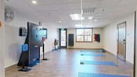 The Wellness Room at 500 Apex Dr in Windsor, Colorado is a thoughtfully designed fitness and movement studio offering a well-rounded space for exercise and wellness activities. The room features durable light wood-tone flooring throughout, a drop tile ceiling with recessed lighting, and clean, neutral-toned walls that create a bright and functional environment. Multiple yoga and exercise mats are laid out across the open floor area, providing ample space for individual or group workouts. The room is equipped with a variety of fitness tools, including hand weights, resistance bands organized on a wall-mounted rack, a BOSU balance trainer, and a colorful medicine ball rack. A SMARTFIT interactive training system panel is mounted prominently along one wall, alongside a wall-mounted flat-screen television. A full-length mirror near the entry allows for form monitoring during exercise. Natural light enters through a window overlooking the surrounding neighborhood, complementing the overhead recessed lighting. Two sets of doors — one wood-paneled entry door and a set of double glass doors — provide convenient access. The space is rounded out with practical amenities including a wall clock, a water station near the exit, and a well-organized resistance band storage area, making this wellness room a comprehensive facility for fitness and recovery.