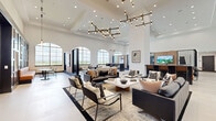 Located at 285 Uptown Blvd in Altamonte Springs, FL, this sophisticated community lounge offers an expansive, single-floor open layout designed with modern elegance. Soaring ceilings define the space, amplified by a series of striking multi-arm branching chandeliers that serve as dramatic focal points throughout. Large arched windows flood the interior with abundant natural light while framing sweeping outdoor views of the surrounding landscape.

The floor plan is thoughtfully organized into distinct functional zones. The central lounge area features richly upholstered leather sofas and accent chairs arranged around a low coffee table, anchored by a bold patterned area rug. Decorative laser-cut partition screens and vertical wood slat wall panels artfully divide the space while maintaining an open, airy feel.

A long, built-in banquette with cognac leather seating lines one wall, complemented by bistro-style tables and chairs beneath tall gridded windows — ideal for casual gathering. A full-length kitchen island with bar seating, warm wood cabinetry, and a herringbone tile backsplash provides a functional hospitality hub. Additional dining areas with sleek black furniture offer versatile seating options. Light-toned large-format tile flooring ties the entire space together, contributing to the clean, contemporary aesthetic.