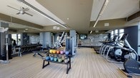 This single-floor fitness center is designed to provide a comprehensive workout environment for users of all fitness levels. The space features light wood-style flooring that extends throughout, lending a clean and energetic feel to the interior. Recessed linear lighting runs across the ceiling, ensuring the entire floor is well-lit and functional.

The strength training area is prominently featured, housing a full rack of dumbbells, a colorful set of kettlebells in graduated weights displayed on a dedicated stand, and multiple weight benches. Several multi-function cable and resistance machines are positioned throughout the space, offering versatility for a wide range of exercise routines.

Along the perimeter, a row of cardio machines — including treadmills and ellipticals — lines the wall, providing ample options for aerobic training. Structural columns are integrated into the layout without impeding traffic flow or equipment access.

A standout design element is the bold, motivational typography mural that covers a large portion of the back wall, featuring fitness-inspired phrases and graphics that contribute to an energizing atmosphere. The combination of functional equipment, thoughtful layout, and dynamic décor makes this fitness center both practical and visually engaging.
