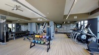 This single-floor fitness center is designed to provide a comprehensive workout environment for users of all fitness levels. The space features light wood-style flooring that extends throughout, lending a clean and energetic feel to the interior. Recessed linear lighting runs across the ceiling, ensuring the entire floor is well-lit and functional.

The strength training area is prominently featured, housing a full rack of dumbbells, a colorful set of kettlebells in graduated weights displayed on a dedicated stand, and multiple weight benches. Several multi-function cable and resistance machines are positioned throughout the space, offering versatility for a wide range of exercise routines.

Along the perimeter, a row of cardio machines — including treadmills and ellipticals — lines the wall, providing ample options for aerobic training. Structural columns are integrated into the layout without impeding traffic flow or equipment access.

A standout design element is the bold, motivational typography mural that covers a large portion of the back wall, featuring fitness-inspired phrases and graphics that contribute to an energizing atmosphere. The combination of functional equipment, thoughtful layout, and dynamic décor makes this fitness center both practical and visually engaging.