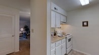 This single-floor residence offers a practical and well-organized layout designed for comfortable everyday living. The kitchen is a standout feature, appointed with crisp white cabinetry that extends from counter height to the ceiling, maximizing storage space. Under-cabinet ventilation sits above a white electric range, complemented by light-toned countertops that provide ample workspace. The cabinetry is fitted with classic hardware, lending a clean and timeless aesthetic throughout the kitchen. A textured ceiling adds subtle architectural character to the space. The kitchen opens directly into the living area, creating a connected flow between the two spaces. The living room, visible through the passageway, is warmly lit by a table lamp and features soft, neutral-toned carpeting. A large indoor plant adds a touch of greenery and life to the room, while a cozy seating arrangement contributes to the inviting ambiance. Together, the spaces create a cohesive, functional interior that balances practicality with a warm, homey atmosphere suited to daily living.