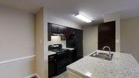 This single-floor space centers around a functional and stylish kitchen that serves as its defining feature. The kitchen is outfitted with rich, dark espresso-toned cabinetry that extends to the upper walls, providing ample storage. Black appliances, including a freestanding gas range with overhead ventilation and a full-sized refrigerator, complement the cabinetry for a cohesive, contemporary look. The light-toned granite countertops offer generous prep space and introduce a subtle contrast against the darker elements. A kitchen island or peninsula with matching granite extends toward the foreground, enhancing both workspace and casual dining potential. A double-basin stainless steel sink with a modern arc-style faucet is integrated into the counter, adding practicality. Overhead, a flush-mounted fluorescent light fixture provides bright, even illumination throughout the kitchen. The neutral beige walls and light-toned flooring contribute to an open, airy feel. A doorway visible in the background hints at additional rooms within the space. Overall, the kitchen presents a clean, well-organized environment suited for everyday living.