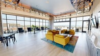 This sophisticated rooftop amenity space is a masterclass in modern design, offering a generously proportioned open-plan layout flooded with natural light through dramatic floor-to-ceiling curtain-wall glazing. Panoramic views of the surrounding waterfront and cityscape are visible from nearly every vantage point, creating a seamless connection between the interior and the outdoors. Wide sliding glass doors open directly onto a spacious terrace furnished with outdoor seating, extending the usable space and blurring the boundary between inside and out.

Inside, the interior design balances warmth and contemporary sophistication. Structural columns clad in stacked natural stone tile add organic texture and visual anchoring throughout the open floor plan. Light hardwood flooring runs continuously underfoot, complementing the airy, neutral palette. A standout feature is the ornate laser-cut metal ceiling installation with a branching, organic pattern that serves as a striking artistic focal point above the lounge seating area.

The space is organized into distinct zones: a comfortable lounge area furnished with vibrant mustard-yellow upholstered chairs and a colorful area rug, a dining and gathering area with modern black tables and chairs, and a sleek bar with a long white countertop and cushioned stools positioned directly in front of a wall of windows. Dual flat-screen displays are mounted on a stone-clad feature wall, rounding out this versatile and visually compelling amenity space.