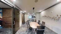 This single-floor professional meeting space is thoughtfully designed to accommodate collaborative work and formal gatherings. At its center sits a smooth oval conference table in a warm wood finish, surrounded by a set of ergonomic high-back chairs upholstered in dark brown, offering both comfort and a refined aesthetic during extended meetings.

Overhead, a striking cluster of matte black pendant lights hangs directly above the table, providing focused task lighting while contributing to the room's contemporary character. Recessed ceiling lights complement the pendants, ensuring the space is evenly and adequately illuminated throughout.

The room is framed by neutral walls accented with subtle crown molding, and the flooring features a richly patterned dark stone or marble-effect surface that adds depth and sophistication. A decorative wall-mounted accent piece on one side adds a artistic touch to the otherwise clean, professional interior.

Sliding glass doors with sleek hardware define the entry, offering visual transparency while maintaining acoustic separation from adjacent areas. Beyond the doors, a glimpse of additional spaces suggests a broader office or amenity environment. The overall ambiance is polished and functional, making this conference room well-suited for business meetings, presentations, or team discussions.