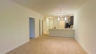 This single-floor residence offers a well-designed open-concept layout that seamlessly blends the living and kitchen areas into one cohesive, functional space. The interior is finished with warm, neutral tones throughout, complemented by plush wall-to-wall carpeting in the main living area that adds comfort underfoot.

The kitchen serves as a focal point of the space, featuring a large peninsula with a clean white countertop that provides ample prep and dining space. Dark cabinetry offers a striking contrast against the light walls, while stainless steel appliances add a modern touch. A stylish pendant light fixture with multiple hanging lights illuminates the kitchen area, enhancing both form and function.

The open living area extends generously from the kitchen, offering flexible space for a variety of furniture arrangements. Neutral cream-colored walls and consistent recessed and overhead lighting throughout create a bright and welcoming ambiance.

Multiple doorways are visible from the main living area, suggesting easy access to additional rooms and hallways. The overall design emphasizes clean lines, a neutral palette, and practical flow between spaces, making this a versatile and comfortable single-level home.