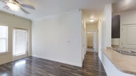 This single-floor space presents a clean, modern aesthetic defined by crisp white walls and rich, dark hardwood-style flooring that runs throughout. The main area opens into a well-lit hallway, illuminated by a ceiling-mounted light fixture, creating a welcoming and functional flow between spaces. The hallway connects to multiple rooms, visible through white-paneled doors, suggesting a thoughtfully organized floor plan. The white walls are smooth and well-finished, complemented by clean white baseboards that provide a polished contrast to the darker flooring. A thermostat and electrical outlets are conveniently positioned along the walls, adding to the practical design of the space. A built-in ledge or shelf is visible along one wall, offering additional surface utility. The neutral color palette throughout creates a bright, airy atmosphere, while the consistent flooring material unifies the space visually. The layout appears open and efficient, with clear sightlines extending through the corridor toward what appears to be carpeted rooms at the far end, providing a subtle variation in texture and comfort across the space.