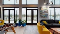 Located at 2200 W Meeker St in Kent, Washington, this single-floor community lounge is a thoughtfully designed social space that blends modern industrial aesthetics with lively, functional amenities. The open-concept layout is anchored by dramatic soaring ceilings with an exposed dark ceiling treatment, from which clusters of Edison-style pendant lights hang at varying heights, creating a warm, inviting atmosphere.

Expansive floor-to-ceiling windows and wide glass-panel doors line the perimeter, flooding the interior with abundant natural light and offering panoramic outdoor views of the surrounding Kent landscape. These doors open directly onto an outdoor terrace, seamlessly connecting the interior to the exterior.

The main gathering area features light wood-look flooring and serves as a versatile social hub, housing a foosball table, a pool table, and a shuffleboard table. Comfortable seating clusters — including a dark sectional sofa and vibrant yellow accent chairs — provide relaxed conversation areas, while bold, colorful artwork adorns the walls throughout.

A well-appointed kitchenette with warm wood cabinetry, stainless steel countertops, a sink, beverage refrigerator, microwave, open shelving, and a large wall-mounted television rounds out the space, making it fully equipped for entertaining and community events.