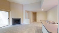 This single-floor space presents an open and functional layout with a clean, neutral aesthetic. The main living area is anchored by a tile-surround fireplace, which serves as a focal point and adds warmth to the room. Soft, neutral-toned walls complement the plush wall-to-wall carpeting, creating a cohesive and comfortable environment throughout.

The space benefits from elevated ceilings in the living area, lending a sense of openness and airiness. A half-wall partition gently defines the transition between the living area and adjacent spaces, maintaining an open flow while offering subtle spatial definition. Recessed lighting provides functional illumination, enhancing the overall brightness of the interior.

A partial view of a countertop along one edge suggests a kitchen or bar area nearby, adding convenience to the layout. Doorways visible in the background lead to additional rooms, indicating a well-connected floor plan. The overall design emphasizes simplicity and practicality, with finishes that provide a neutral canvas well-suited for a variety of interior styles.