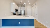 This thoughtfully designed single-floor residence combines modern aesthetics with functional living. The kitchen is the undeniable centerpiece, showcasing a striking two-tone design with bold blue lower cabinetry paired against crisp white upper cabinets. A large island with a white quartz countertop and an undermount sink serves as both a workspace and a gathering point, illuminated by stylish pendant lights. A decorative geometric tile backsplash adds visual interest, while a full suite of stainless steel appliances — including a refrigerator, range, microwave, and dishwasher — provides a polished, professional finish. Light-toned hardwood flooring flows consistently throughout the space, creating a warm, cohesive feel. The bedroom is bright and airy, benefiting from a generously sized window that invites natural light and offers views of lush greenery outside. An adjacent room features built-in shelving, ideal for use as a closet or storage area. The bathroom is well-appointed with a granite-topped navy vanity and is conveniently located adjacent to a dedicated laundry closet housing a stacked washer and dryer unit. Neutral warm-gray walls throughout complement the contemporary finishes, creating a clean, inviting ambiance across the entire residence.