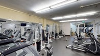 This fitness center offers a fully equipped workout environment designed to accommodate a wide variety of exercise needs. The space is laid out efficiently across a single floor, with equipment organized to allow comfortable movement between stations. Strength training is well represented, with multiple cable machines and weight systems—including selectorized stack machines and free weights—arranged along mirrored walls that enhance the sense of depth and allow users to monitor their form. Cardio equipment, including a Matrix treadmill, is positioned for easy access. The flooring features durable, dark rubberized material suited for heavy use and providing cushioning underfoot. Overhead, bright fluorescent lighting ensures the entire space is evenly and thoroughly illuminated. The walls carry a light, neutral tone that keeps the environment feeling open and functional. A secondary area visible through a doorway at the rear of the space suggests additional fitness or utility zones. Overall, the fitness center is clean, practical, and thoughtfully arranged to serve the diverse training goals of its users.