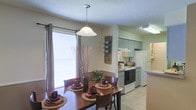 This single-floor residence showcases an open-concept layout that seamlessly connects the dining area and kitchen, creating a cohesive and functional living space. The dining area is a welcoming focal point, highlighted by a classic pendant light suspended above a wooden dining table with seating for multiple guests. A large window fitted with horizontal blinds and curtain panels allows generous natural light to fill the room, enhancing the warm, neutral color palette of the walls and flooring.

The kitchen is efficiently arranged and accessible directly from the dining space, featuring light gray cabinetry that extends to the upper walls for ample storage. White appliances, including a freestanding range and a full-size refrigerator, provide a clean, consistent look. A range hood is positioned above the stove, and under-cabinet lighting adds practicality to the workspace. Light-toned flooring runs throughout the kitchen, contrasting softly with the cabinetry.

The textured ceilings are consistent throughout both areas, contributing to the overall cohesive aesthetic. Decorative touches, including wall-mounted accents and tabletop arrangements, add character to the space, making it feel both livable and inviting.