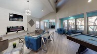 This clubhouse is a well-appointed community gathering space designed with modern aesthetics and a welcoming atmosphere. The open floor plan flows seamlessly across light hardwood-style flooring, unifying multiple distinct activity zones under soaring vaulted ceilings. A striking sputnik chandelier anchors the main lounge area, which features a shiplap-clad fireplace with a mounted flat-screen television above and a comfortable seating arrangement. Teal accent walls and bold blue velvet seating add vibrant pops of color against the neutral grey and white palette throughout.

Adjacent to the lounge, a game area provides a full-sized billiards table beneath a modern geometric pendant light, with large floor-to-ceiling windows offering views of the outdoor pool area and filling the space with natural light. Tufted blue accent chairs are arranged along the windows, creating an inviting nook for relaxation.

The clubhouse also includes a compact kitchenette outfitted with dark cabinetry, light quartz countertops, a stainless steel refrigerator, and shiplap accent walls — ideal for hosting community events. A dining area with teal-painted walls and ample natural light rounds out the space. Decorative wall art and carefully curated furnishings throughout contribute to a cohesive, contemporary design that is both functional and visually engaging.