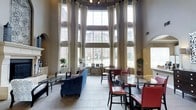 This impressive single-floor space makes an immediate statement with its dramatic, floor-to-ceiling windows that stretch to extraordinary heights, flooding the interior with abundant natural light and offering clear views of the surrounding exterior. Tall, flowing drapes frame the expansive window arrangement, adding an element of refined elegance to the grandeur of the architecture.

The space is thoughtfully organized into distinct seating zones. A relaxed lounge area features a plush blue sofa and patterned accent chairs arranged around a sleek glass coffee table, creating an inviting setting for casual conversation or relaxation. Adjacent to this, a dining area is furnished with round glass-top tables paired with rich red leather chairs and dark wood seating, providing a more structured environment for gatherings or collaborative use.

Overhead, a decorative chandelier anchors the ceiling and complements the room's sophisticated aesthetic. The flooring throughout is composed of large-format neutral tiles, contributing to the open and airy feel of the space. Arched architectural details are visible along the walls, adding classical character. Potted greenery is thoughtfully placed to introduce natural warmth. Overall, the space combines architectural grandeur with comfortable, functional furnishings to create a welcoming and visually striking communal environment.