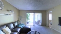 This single-floor space centers around a comfortable living room characterized by neutral-toned walls and plush wall-to-wall carpeting that adds warmth underfoot. The room's most notable feature is a large sliding glass door fitted with vertical blinds, which serves as both a source of abundant natural light and a direct connection to an outdoor patio. Through the glass, potted tropical plants and surrounding greenery are clearly visible, lending the interior a refreshing, nature-adjacent atmosphere. The ceiling features a textured popcorn finish, a classic detail that adds dimension overhead. The living area is generously proportioned, comfortably accommodating a sectional leather sofa and a glass-top coffee table, with additional space for decorative elements such as ornamental branches and accent plants. The overall ambiance is relaxed and inviting, enhanced by the bright outdoor light that filters through the sliding door, making the space feel open and airy despite its single-floor layout. The seamless indoor-outdoor connection is a defining characteristic of this living space.