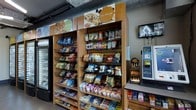 This single-floor retail space functions as a compact convenience or micro-market area, designed to provide quick access to a wide variety of snack and beverage options. The layout centers around tall, warm-toned wooden shelving units that line the walls and display an extensive selection of packaged goods, including chips, nuts, granola bars, instant noodles, and other grab-and-go items. Overhead signage, including branded labels such as "Recharge" and "Replenish," helps organize the merchandise into themed categories, guiding shoppers toward relevant product types efficiently. Wire basket inserts on the shelves add a functional, organized aesthetic while maximizing product visibility. Along the right side of the space, a self-service Avenue kiosk provides a modern, automated checkout solution, streamlining the purchasing process. A wall-mounted monitor is also visible, likely used for informational or promotional display purposes. A refrigerated unit along the left wall offers chilled beverage options. The overall ambiance is utilitarian and efficient, with gray industrial-style ceilings and neutral flooring contributing to a clean, no-frills environment well-suited for high-traffic locations such as office buildings, campuses, or transit hubs.