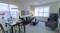 This single-floor residence offers an open-concept layout that seamlessly connects the dining and living areas, creating a cohesive and functional living environment. The living room is anchored by a dark charcoal sofa accented with decorative pillows in earthy tones, complemented by a rectangular coffee table with a lower storage shelf. A cushioned accent chair adds additional seating, while a table lamp provides warm ambient lighting. A large window with horizontal blinds allows generous natural light to fill the space, overlooking greenery outside.

The dining area sits adjacent to the living room and features a dark-finished table paired with cushioned chairs in a sage green upholstery, lending a pop of color to the neutral palette. A pendant chandelier hangs above the dining table, adding a decorative focal point. The dining area also benefits from its own window, further brightening the space.

The walls are painted in a soft, neutral tone, and light-colored carpeting runs throughout, contributing to the overall airy and welcoming ambiance. A trio of framed artwork on the shared wall between the two areas ties the spaces together visually, making this a well-appointed and comfortable home.