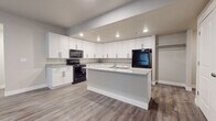 This single-floor unit offers a clean, contemporary design highlighted by a cohesive neutral color palette and wood-look luxury vinyl plank flooring throughout the main living areas. The kitchen serves as a central feature, showcasing white shaker-style cabinetry extending to the ceiling for generous storage, light granite countertops, and a matching center island with a built-in sink. Black stainless appliances, including a range, microwave, and refrigerator, provide a modern contrast against the bright cabinetry, while recessed lighting ensures the space is well-lit. The living area transitions seamlessly from the kitchen, featuring a glazed entry door that allows natural light to filter in. Bedrooms are carpeted for comfort and receive natural light through window treatments. A full bathroom features white cabinetry with granite countertops, a mirrored vanity, and a shower-tub combination. The dedicated laundry room is a practical highlight, offering side-by-side washer and dryer units, additional white cabinetry with granite countertops, and a small window for ventilation. Hallways connect the rooms efficiently, with closet spaces providing organized storage throughout.