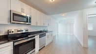 This well-appointed single-floor unit offers a thoughtfully designed open-concept layout that seamlessly connects the kitchen and living area. The kitchen is a standout feature, outfitted with crisp white shaker-style cabinetry, dark granite countertops, and a full suite of stainless steel appliances including a refrigerator, electric range, over-the-range microwave, and dishwasher. Light wood-style flooring runs throughout the main living areas, complementing the bright, neutral color palette and giving the space a clean, modern aesthetic. The open living area is generously proportioned, with large vertical blinds allowing natural light to filter in while maintaining privacy. The bedroom is a calm, minimalist space featuring the same light hardwood flooring, white walls, and a window with blinds for natural light. The bathroom is finished with large-format beige floor tiles, grey stone-look tile surrounding the soaking tub, and clean white fixtures. Flush-mount ceiling lighting throughout keeps the overall look uniform and uncluttered. The layout, as visible in the dollhouse view, is efficient and well-organized, making excellent use of the available footprint for comfortable everyday living.