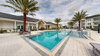 Located at 1520 Alexis Ave in Daytona Beach, FL, this fitness center offers a comprehensive workout environment spread across a single, generously sized floor. The main exercise room is a standout space, featuring striking geometric-patterned carpet in shades of blue, gray, and white that lends a dynamic, modern aesthetic. Ceiling fans and recessed lighting keep the area bright and comfortable, while a row of large windows along the walls floods the space with natural light and views of the surrounding landscape. The main floor is outfitted with an extensive array of equipment, including treadmills, cable machines, a multi-functional power rack, a punching bag, TRX suspension trainers, a plyo box, weight benches, and a full dumbbell rack — catering to cardio, strength, and functional training alike. A mounted flat-screen TV adds convenience for entertainment during workouts. Adjacent to the main floor, a separate studio room provides a quieter setting with light hardwood-style flooring, full-length mirrors, a wall-mounted TV, and equipment such as stationary bikes, a rowing machine, and stability balls — ideal for low-impact or focused training sessions. The hallway connects the two spaces seamlessly, completing this well-appointed, full-service fitness facility.