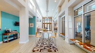 This single-floor community space makes a striking first impression with its grand entrance — a floor-to-ceiling wood-framed glass door surround that floods the interior with natural light and frames outdoor views beautifully. The soaring ceilings, supported by structural columns and clerestory windows along the upper walls, lend an airy, open atmosphere throughout.

Light-toned wood-look tile flooring runs seamlessly across the space, complementing the crisp white walls accented by bold teal feature walls and warm neutral tones. The central gathering area features a round bistro-height table with modern bentwood chairs, set atop a geometric patchwork area rug, creating an inviting hub for casual interaction.

Adjacent to the main entry, a relaxed seating nook offers a teal mid-century modern sofa, a sleek white coffee table, and a patchwork hide rug beneath large windows that invite natural light. Further into the space, a dedicated lounge area showcases a stacked-stone fireplace with a wall-mounted television, flanked by rustic wood accent walls and furnished with white lounge chairs and eclectic side tables. A large-scale connect-four game adds a playful, social touch to the room. The overall ambiance is modern, welcoming, and thoughtfully designed for both comfort and community engagement.