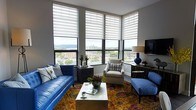 This thoughtfully designed single-floor space makes a striking impression through its architectural use of large, floor-to-ceiling corner windows that flood the interior with natural light and frame panoramic views of the surrounding cityscape and distant mountain ridgeline. The living area serves as the centerpiece of the space, showcasing a bold blue leather sofa paired with a classic neutral armchair and a complementary blue accent chair, all arranged around a sleek coffee table. Underfoot, a richly patterned area rug in warm jewel tones anchors the seating arrangement and adds a vibrant, artistic energy to the room. The corner window configuration creates an open, expansive feel, drawing the eye outward to the urban landscape below. Layered roller shades offer flexible light control while maintaining the connection to the outside. A wall-mounted television and coordinating media console provide functional convenience, while decorative elements — including sculptural accents, a cobalt blue table lamp, and curated accessories — lend personality and polish to the space. The overall ambiance balances contemporary design with warm, inviting details, resulting in a space that feels both sophisticated and livable.
